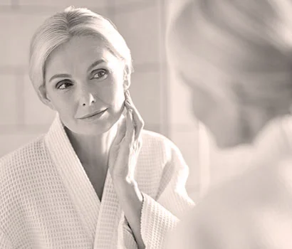 50 years old blonde woman standing in bathroom after shower touching face, looking at reflection in mirror